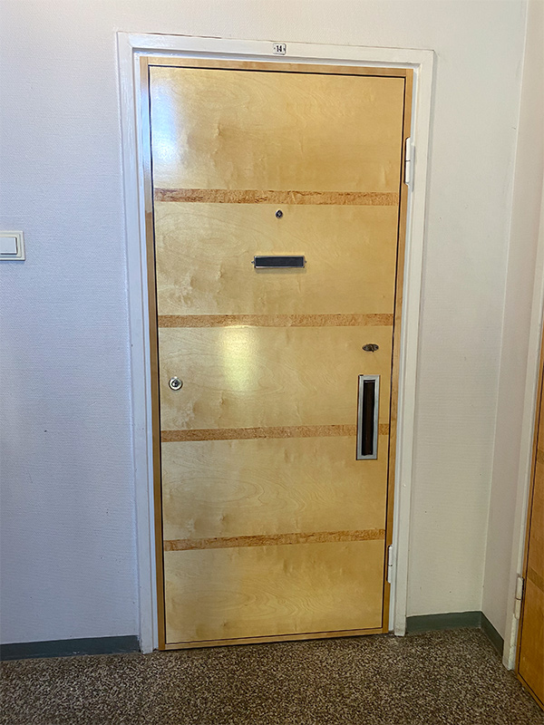 Valokuva rappukäytävästä suljetusta kerrostason puisesta ovesta. Ovessa on vaakasuuntaiset koristelistat, ovisilmä, avaimenreikä ja postiluukku.